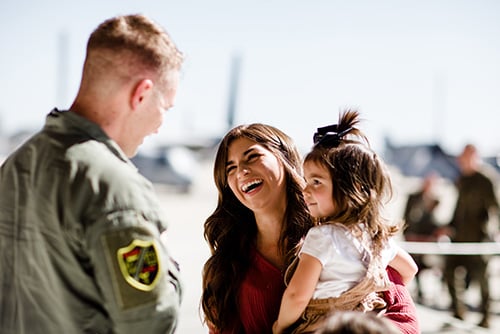 Marine reuniting with his proud wife, while she laughs and holds their toddler daughter on her hip