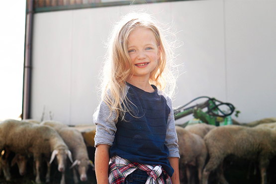 Young blond girl in sunlight, smiling in front of a herd of sheep on her family's farm near Dickinson, ND