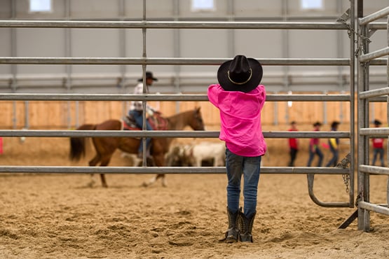 Young man in a magenta shirt, leaning against a cattle gate as he watches cowboys warm up at the Jamestown rodeo